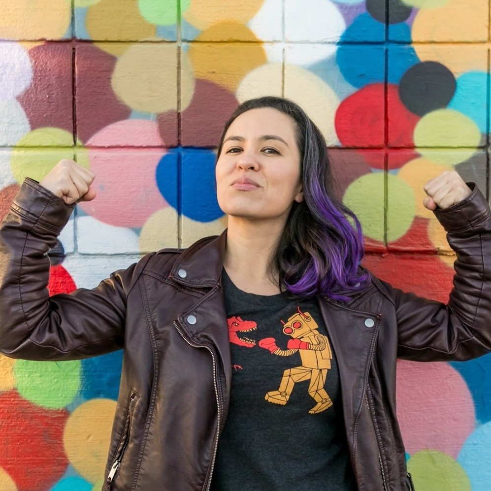 A woman in black and purple hair and a black leather jacket flexes her biceps in front of a brightly colored wall.