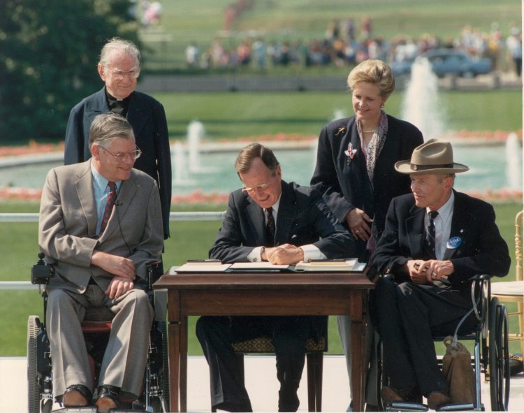 President George HW Bush sits at a table outside in the sun and signs a document.  He is flanked on either side by two men in suits who are in wheelchairs.  Two people watch.  In the distance behind them is a fountain and a green meadow.