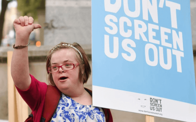 Heidi Crowter raising her fist and holding a Don't Screen Us Out placard